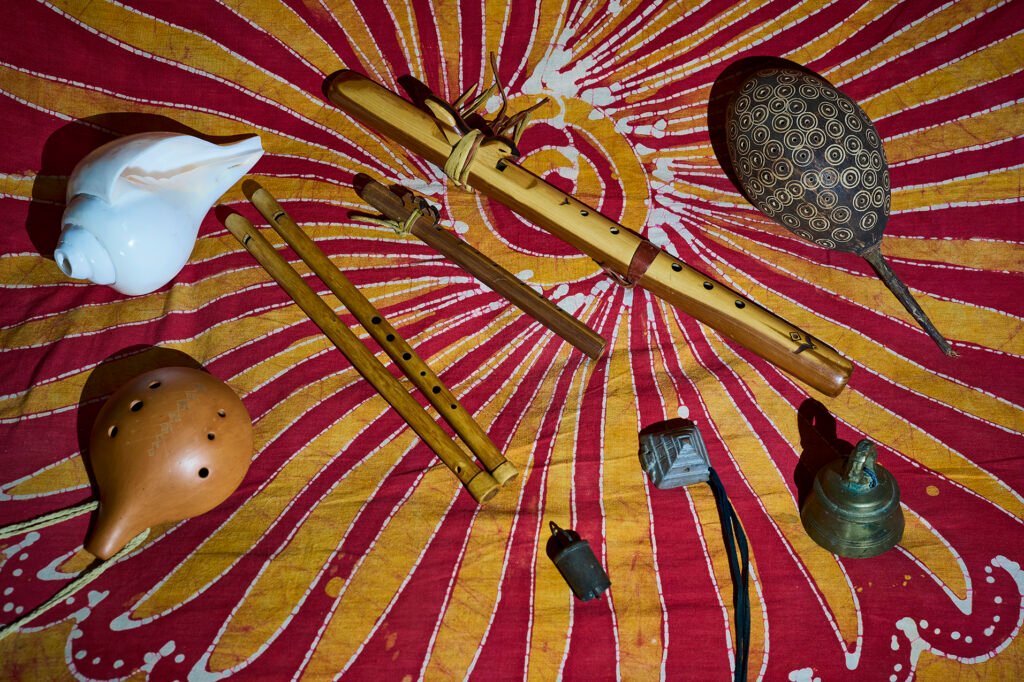 Small resonant instruments laid out on a red and gold cloth: conch shell, ocarina, bamboo flutes, rattle and bells